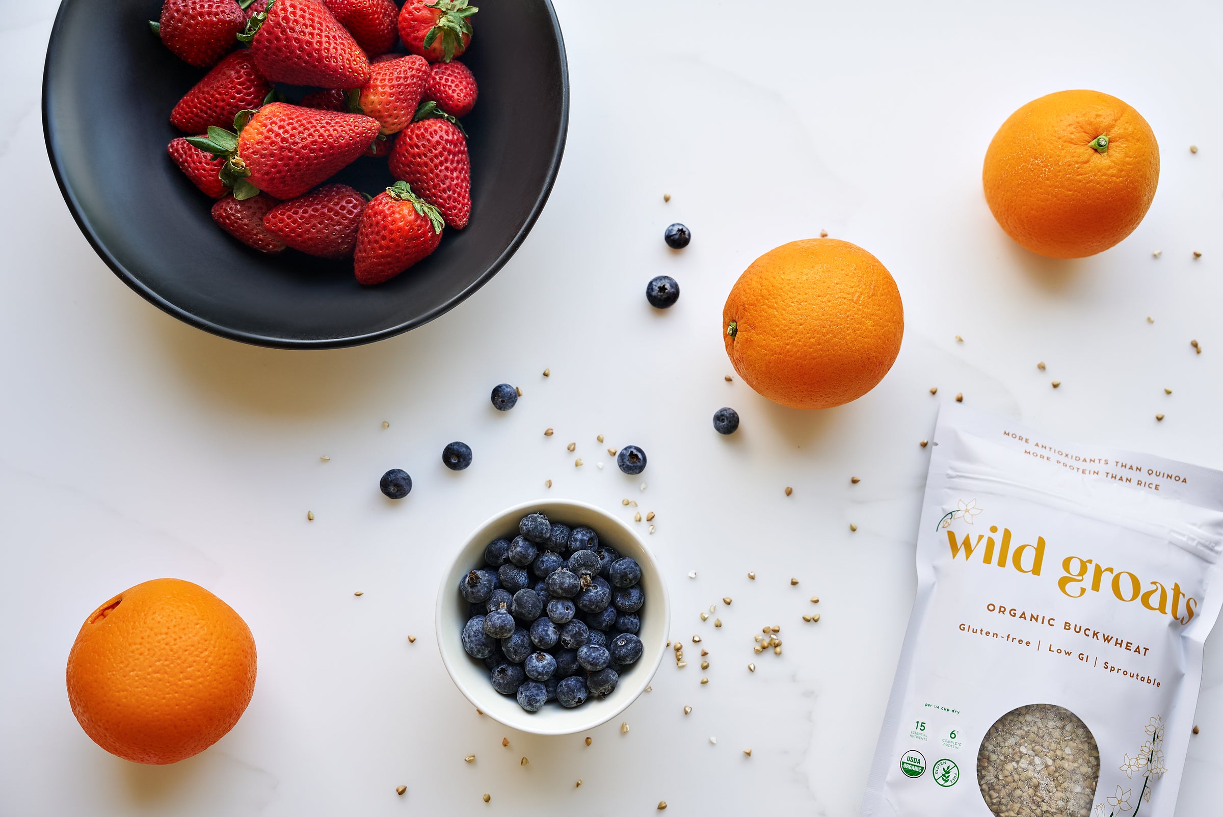 Wild Groats bag with fresh strawberries, oranges, blueberries and loose buckwheat groats on white countertop
